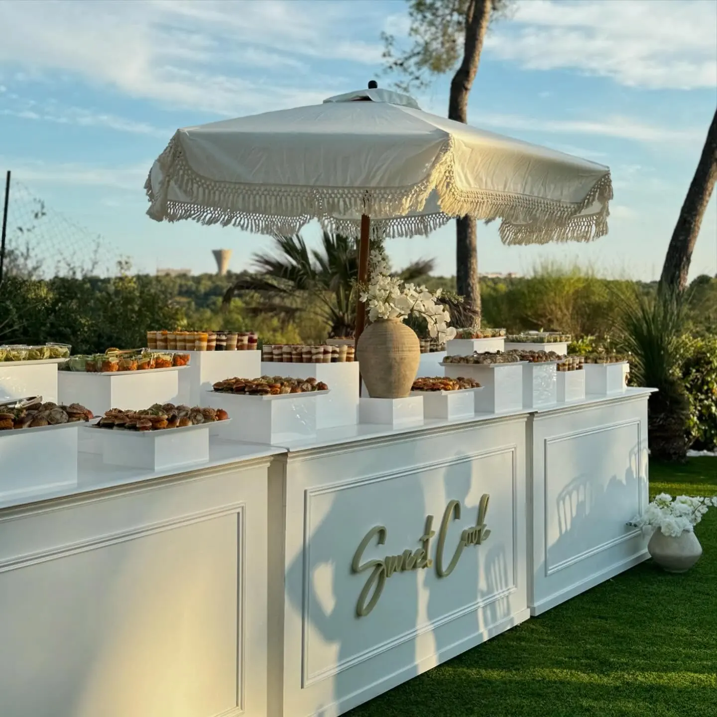 Buffet Sweet Cook en extérieur au coucher du soleil : comptoirs blancs, présentoirs de mignardises et parasol à franges